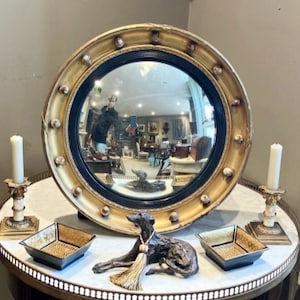 Könnte beinhalten: Ein runder, goldgerahmter Spiegel mit einem schwarzen Innenring, der einen Raum reflektiert. Der Spiegel ist von dekorativen Elementen umgeben, darunter zwei Kerzenhalter mit weißen Kerzen, zwei quadratische Schalen und eine Bronzestatue eines Hundes mit Quaste.