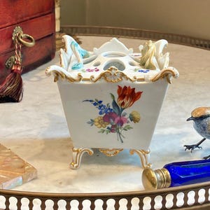 May include: A white porcelain square flower frog or bulb pot with gold-trimmed edges and feet. The exterior features a colorful floral bouquet painting. The top has multiple circular openings and small sculpted decorative elements in blue and tan.