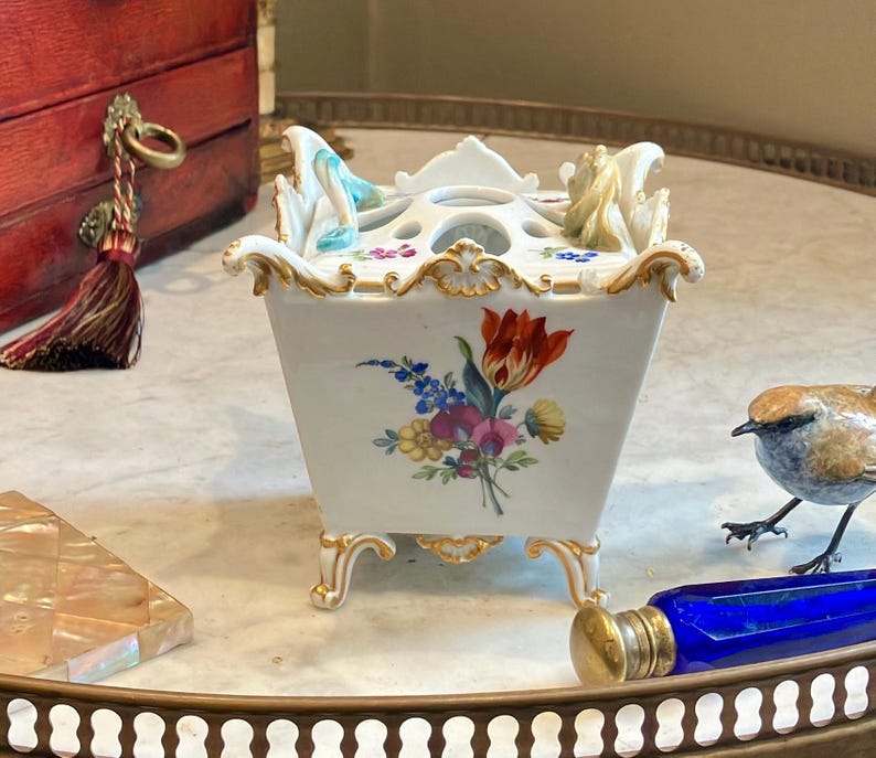 May include: A white porcelain square flower frog or bulb pot with gold-trimmed edges and feet. The exterior features a colorful floral bouquet painting. The top has multiple circular openings and small sculpted decorative elements in blue and tan.