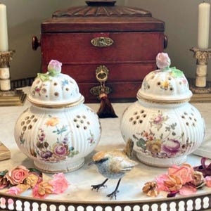 Può includere: Due vasi decorativi in ceramica bianca con disegni floreali e fori traforati, sormontati da un terminale a forma di rosa. Una piccola statuina di uccello è in piedi davanti ai vasi. Sullo sfondo c'è una scatola di legno scuro.