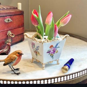 May include: A white porcelain flower vase with a square shape, gold-trimmed edges, and four ornate feet. The vase features hand-painted floral motifs in pink, blue, and yellow, and contains several pink tulips with green stems and leaves.
