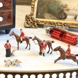 Może przedstawiać: Miniaturowe figurki jeździeckie, w tym jeźdźcy w czerwonych płaszczach na brązowych koniach, kobieta w czerwonej kurtce i małe psy. Widoczna jest również czerwona szklana butelka ze srebrnymi akcentami.