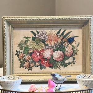 Pode incluir: Uma impressão floral emoldurada com um buquê de flores rosa, vermelhas e brancas com folhagem verde. A moldura dourada tem detalhes ornamentados. Uma figura decorativa de pássaro e duas tigelas pequenas estão em frente à moldura.