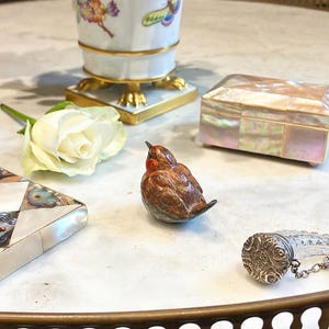 May include: A small, brown and red bird figurine with a white rose and a mother of pearl box on a marble surface.