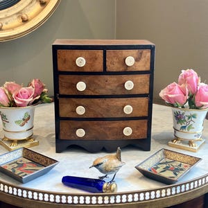 Può includere: Un piccolo comò in legno con cinque cassetti, con finitura a venatura scura e pomelli bianchi. Il comò è posto su un tavolo con piano in marmo, accompagnato da composizioni floreali in vasi decorativi e piccoli vassoi.
