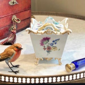 May include: A white porcelain square container with a lid, featuring hand-painted floral motifs in pink, blue, and yellow. The piece has ornate gold-trimmed edges, four curved feet, and a central handle on the lid, showcasing delicate craftsmanship.