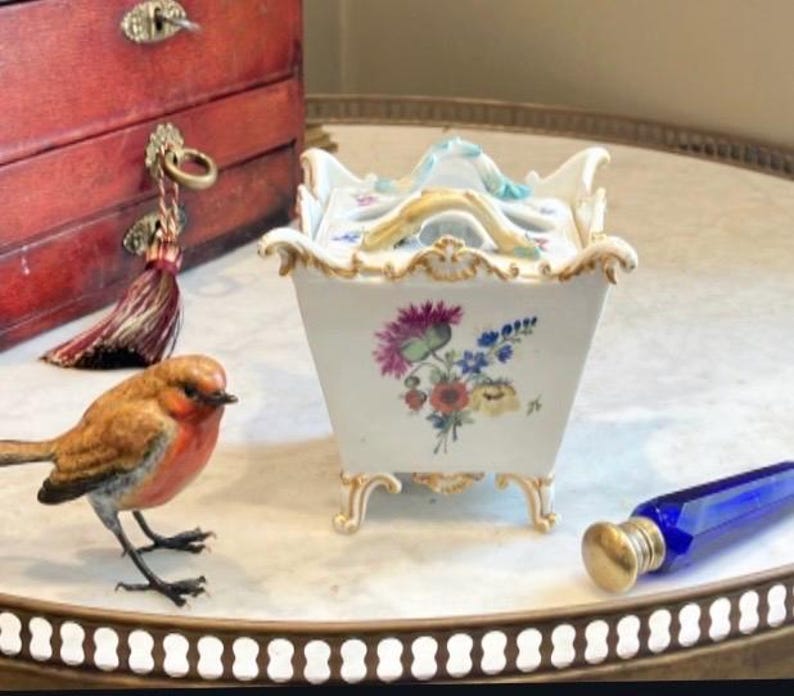 May include: A white porcelain square container with a lid, featuring hand-painted floral motifs in pink, blue, and yellow. The piece has ornate gold-trimmed edges, four curved feet, and a central handle on the lid, showcasing delicate craftsmanship.