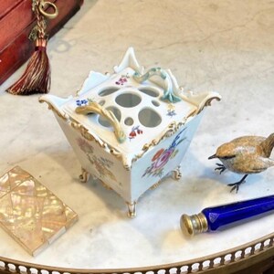 May include: A square white porcelain flower frog with a removable lid featuring multiple circular openings. The vessel is decorated with hand-painted floral motifs, gold trim, and four small feet. Two curved handles extend from the top lid.
