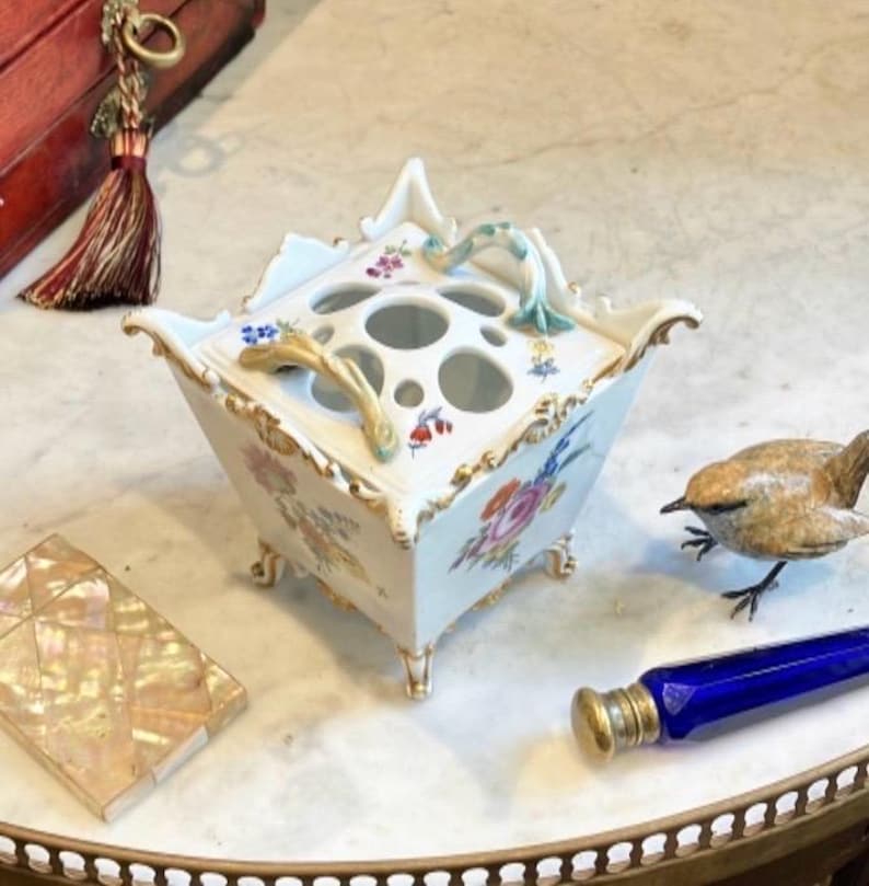 May include: A square white porcelain flower frog with a removable lid featuring multiple circular openings. The vessel is decorated with hand-painted floral motifs, gold trim, and four small feet. Two curved handles extend from the top lid.