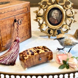 May include: An arrangement of vintage decorative objects on a white surface. Features a wooden box with a tassel, a small tortoiseshell box, a gold framed portrait, a bird figurine, and pink roses.