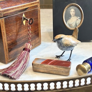May include: A wooden box with a key and a red and gold tassel, a small bird figurine, a rectangular metal box with a banded agate lid, a portrait in a black frame, and a blue glass bottle with a brass stopper arranged on a metal tray.