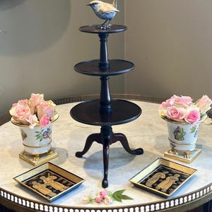 Può includere: Un supporto a tre livelli in legno scuro con una piccola statuetta di uccello sul ripiano superiore, affiancato da due vasi in porcellana bianca pieni di rose rosa e due vassoi rettangolari neri e oro con figure su una superficie di marmo.