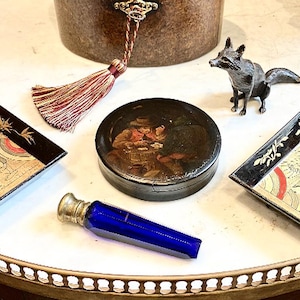 May include: A vintage, round, wooden box with a painted scene of two people in period clothing. The box is closed and has a silver lid. The box is sitting on a white surface with a blue glass bottle stopper and a small silver fox figurine.