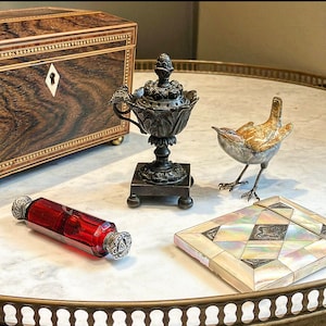 Könnte beinhalten: Eine Sammlung antiker Dekorationsgegenstände auf einer Marmortischplatte. Eine Schachtel aus Silber und Perlmutt, eine kleine rote Glasflasche mit einem silbernen Stopfen, ein Bronzene Räuchergefäß und eine kleine Keramikvogelfigur.