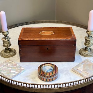 May include: A rectangular wooden box with a central oval inlay, flanked by two brass candlesticks with light pink candles. In front, a small round box features a portrait miniature, accompanied by two square mother-of-pearl inlaid coasters.