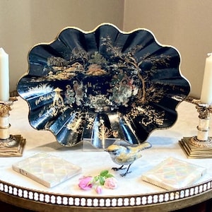May include: A black, scalloped decorative tray with a gold rim and a detailed landscape scene. Two white candles sit in ornate gold candle holders. A small bird figurine, two square mother-of-pearl boxes, and pink flowers complete the display.