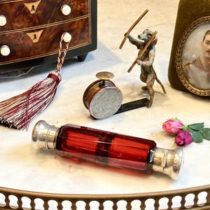 Puede incluir: Una variedad de objetos antiguos sobre una superficie de mármol. Incluye un frasco de perfume de vidrio rojo, un pequeño cofre de madera, un tambor en miniatura, una figura de perro, un retrato enmarcado y una borla.