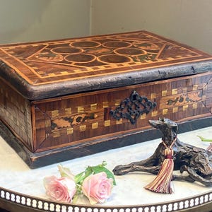 May include: A rectangular wooden box with a decorative inlaid design on the lid and sides. The box is brown with lighter wood accents and a dark border. A bronze dog statue with a tassel and pink roses are in front of the box.