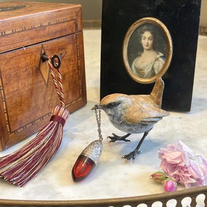 May include: A decorative still life featuring a wooden box, a framed portrait, and a bronze bird figurine. The bird holds a red and silver perfume bottle. A tassel and pink flowers complete the arrangement.