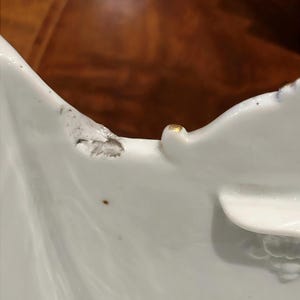 May include: A white porcelain bowl with a scalloped edge featuring a small gold-painted accent on the rim and a noticeable area of gray surface damage or chipping along the upper curved border of the ceramic structure.