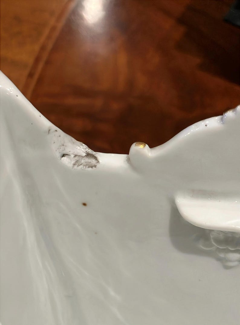 May include: A white porcelain bowl with a scalloped edge featuring a small gold-painted accent on the rim and a noticeable area of gray surface damage or chipping along the upper curved border of the ceramic structure.