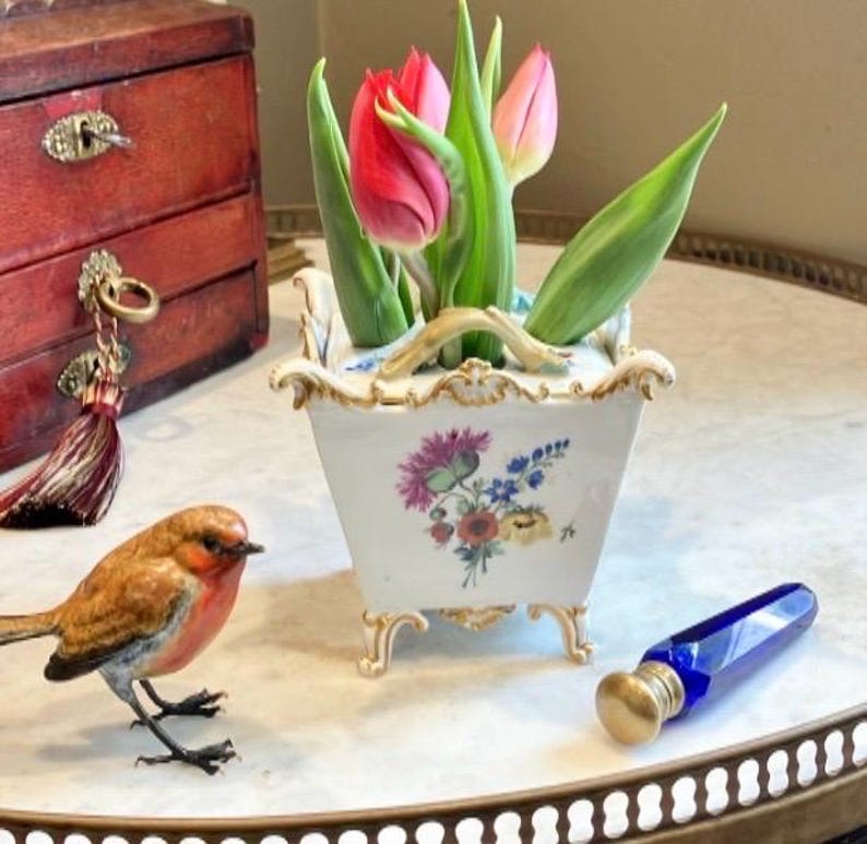 May include: A square white porcelain planter with gold-trimmed edges and feet, featuring a floral bouquet design. It holds three tulips in shades of red and pink. Nearby sits a small brown bird figurine and a cobalt blue glass bottle with a gold cap.