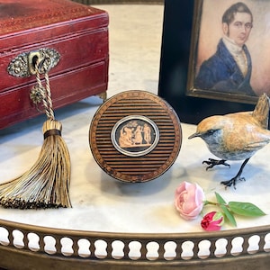 May include: A round wooden box with horizontal black and brown stripes, featuring a central oval medallion with a black and orange classical relief scene. The lid is decorated with a thin, intricate border pattern around the central inlay.
