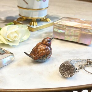 May include: A small, intricately detailed, brown and blue bird figurine with a red head. The bird is sitting on a white marble surface. There is a white rose, a small silver box with a clear glass top, and a decorative box with a mother-of-pearl inlay in the background.