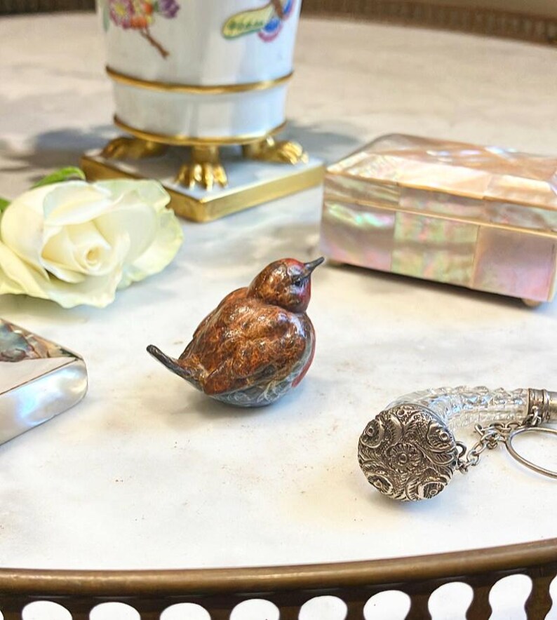 May include: A small, intricately detailed, brown and blue bird figurine with a red head. The bird is sitting on a white marble surface. There is a white rose, a small silver box with a clear glass top, and a decorative box with a mother-of-pearl inlay in the background.
