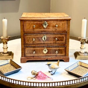 Può includere: Un piccolo cassettone in legno con tre cassetti, ferramenta in ottone e dettagli intarsiati. Due candelabri con candele accese affiancano il cassettone. Vassoi decorativi e una statuina di uccello sono sul tavolo.