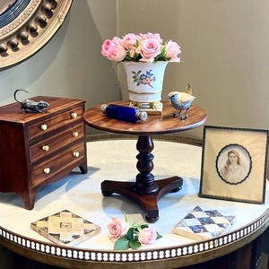 Può includere: Un tavolino rotondo in legno con un vaso di rose rosa, una bottiglia di vetro blu e un ritratto incorniciato. Sono presenti anche un piccolo comò in legno e oggetti decorativi.