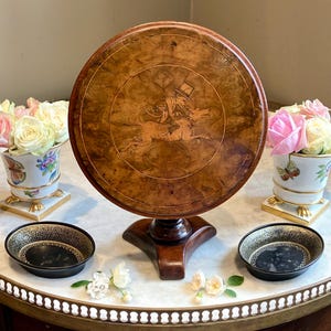 May include: A round wooden table with a central inlay depicting a figure in a top hat riding a dog. The table features a polished burl wood grain finish and stands on a tripod base. Two small black bowls with gold rims and two floral vases are present.