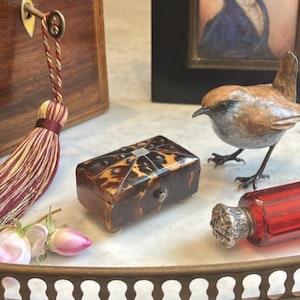 May include: A decorative arrangement featuring a small, tortoise-shell patterned box with silver accents, a bronze bird figurine, and a red glass bottle with a silver stopper. A tassel and pink roses are also present.