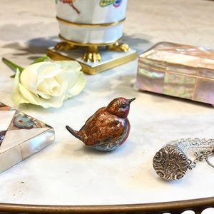 May include: A small, intricately detailed bird figurine made of painted porcelain. The bird is brown, red, and blue with a detailed pattern. The figurine is sitting on a white surface.