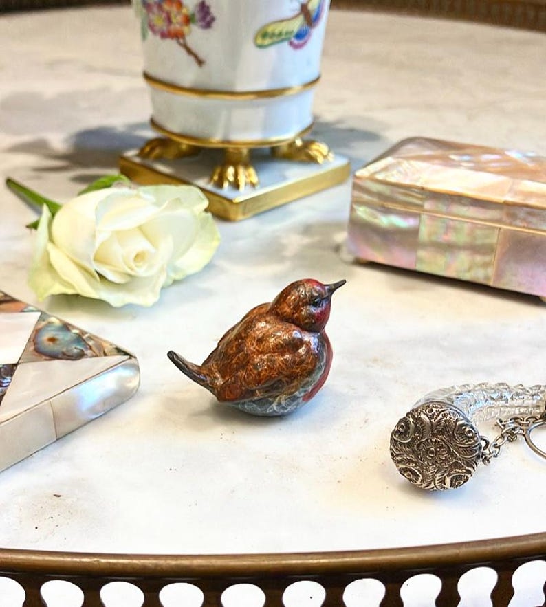 May include: A small, intricately detailed bird figurine made of painted porcelain. The bird is brown, red, and blue with a detailed pattern. The figurine is sitting on a white surface.