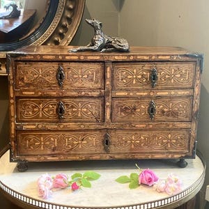 Può includere: Un cassettone in legno a sei cassetti, con intricati disegni intarsiati floreali. Il cassettone è sormontato da una scultura in bronzo di un cane. Il pezzo è posto su un tavolo con piano in marmo, con rose rosa alla base.