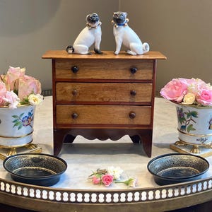 Può includere: Una piccola cassettiera in legno a tre livelli sormontata da due statuette di carlino in ceramica. Ai lati, due vasi in porcellana bianca riempiti di rose rosa, e in primo piano due ciotole nere con motivi in filigrana dorata.