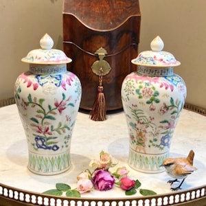 May include: Two ornate ceramic ginger jars with lids, featuring floral designs in pink, blue, and green. They are displayed on a decorative tray with roses and a small bird ornament. A wooden box is in the background.