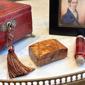 May include: A rectangular wooden box with a polished light brown wood grain finish, featuring dark diagonal inlay lines and a small silver-colored rectangular plaque on the lid. The box has a single front knob and rests on small white feet.
