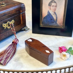 May include: A small, dark wood coffin-shaped box with a heart-shaped inlay on the lid and a diamond-shaped inlay on the side. The box is positioned near a wooden chest with a brass keyhole, a framed portrait of a man, and two small roses.