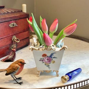 May include: A white porcelain flower vase with gold trim and a floral motif, containing pink tulips. Beside it sits a small brown bird figurine and a cobalt blue glass bottle with a gold cap. The vase features four feet and a multi-hole top.