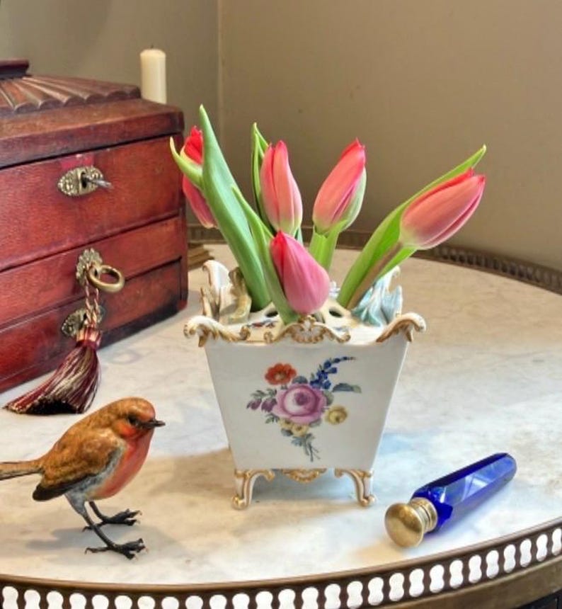 May include: A white porcelain flower vase with gold trim and a floral motif, containing pink tulips. Beside it sits a small brown bird figurine and a cobalt blue glass bottle with a gold cap. The vase features four feet and a multi-hole top.