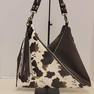 May include: A brown and white cowhide-patterned shoulder bag with a dark brown leather trim and straps. The bag features a zipper closure, a tassel, and gold-colored hardware. The bag is displayed on a black stand.