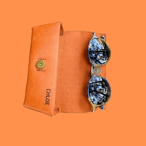 Puede incluir: Un estuche de gafas de sol de cuero marr&oacute;n con el nombre "CHLOE" grabado en la parte delantera. El estuche est&aacute; abierto y un par de gafas de sol redondas con lentes oscuras est&aacute;n dentro.