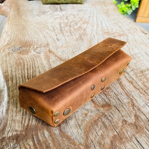 Puede incluir: Una funda de cuero marr&oacute;n con cierre de solapa y remaches met&aacute;licos. La funda tiene la palabra "BOARD" grabada en relieve en la solapa.