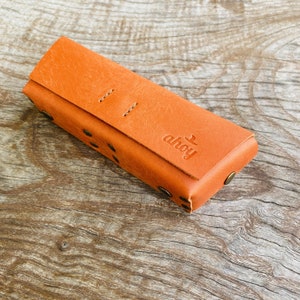 Puede incluir: Una funda de cuero marr&oacute;n con la palabra "ahoy" grabada en la parte delantera. La funda tiene una solapa que se cierra con un broche a presi&oacute;n y est&aacute; decorada con peque&ntilde;os remaches met&aacute;licos.