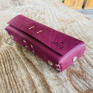 Puede incluir: Una funda de cuero burdeos con tachuelas plateadas y la palabra "ahoy" grabada en la parte delantera.