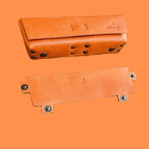 Puede incluir: Una cartera de cuero marr&oacute;n con cierre de solapa y la palabra "dhoy" grabada en la parte delantera. La cartera est&aacute; abierta para revelar un interior de cuero marr&oacute;n con un solo bolsillo. La cartera est&aacute; sobre un fondo naranja s&oacute;lido.