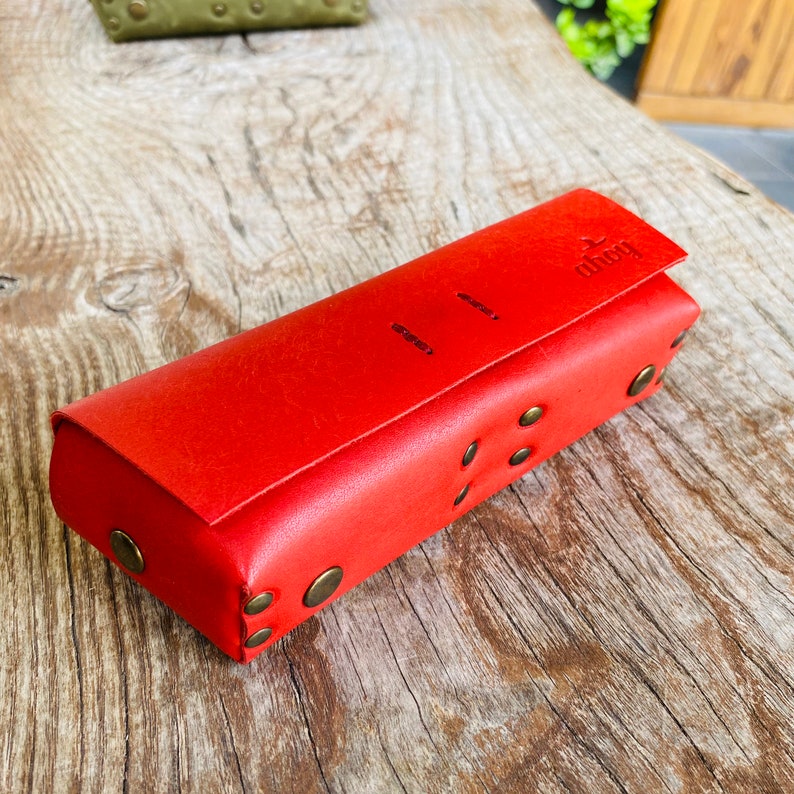 Puede incluir: Una funda de cuero roja con cierre de solapa y la palabra "ahoy" grabada en la parte delantera. La funda tiene remaches decorativos de lat&oacute;n alrededor de los bordes.