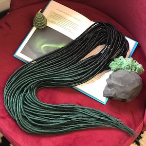 May include: A bundle of black and teal dreadlocks, with a gradient effect, resting on a burgundy velvet chair. A green tree-shaped candle and a concrete skull-shaped planter with greenery are also visible.
